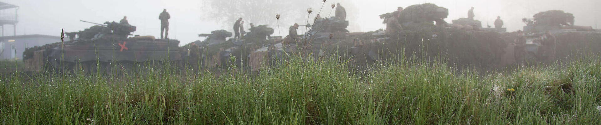 Panzer und Soldaten üben im morgendlichen Nebel hinter einer grünen Wiese während des Gefechtsbild-Trainings.