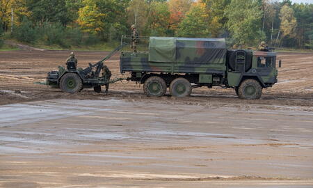 Ein militärischer LKW zieht ein schweres Geschütz durch den schlammigen Boden eines Übungsgeländes im Herbst.