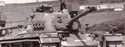 Ein Soldat steht auf einem Panzer in ländlicher Umgebung, während im Hintergrund ein Turm sichtbar ist.