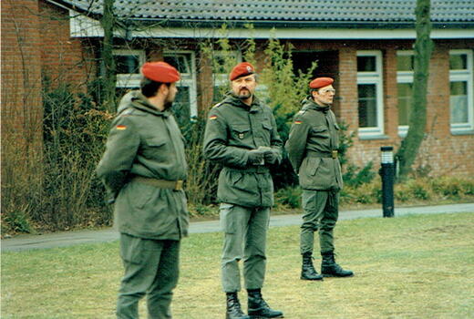 Drei Soldaten in grüner Uniform und roten Baretten stehen auf einem Rasen vor einem Backsteingebäude.