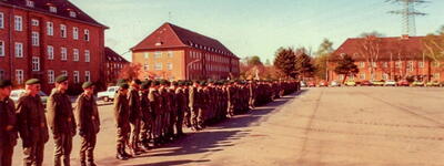 Soldaten stehen in einer Reihe auf einem Kasernenhof in Angetreten, umgeben von roten Backsteingebäuden.