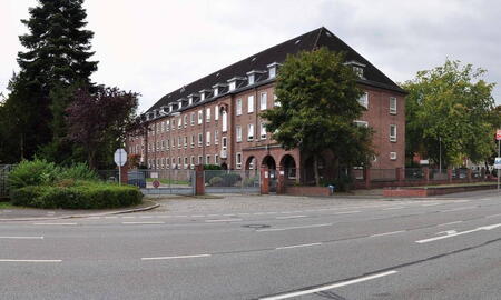 Ein historisches Backsteingebäude der Scholtz-Kaserne von 2008 steht an einer ruhigen Straßeneinfahrt, umgeben von Bäumen.