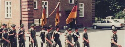 Soldaten marschieren in Formation mit deutschen Flaggen vor einem Backsteingebäude, während ein Auto im Hintergrund steht.