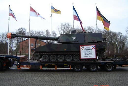 Ein Panzer wird auf einem LKW transportiert, während im Hintergrund mehrere Flaggen im Wind wehen.