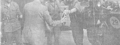 Ein Mann in Anzug überreicht Soldaten in Uniformen ein Getränk neben einem Militärfahrzeug.