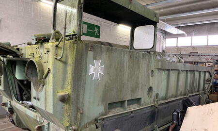Ein alter, moosbedeckter Militärtransporter mit einem Kreuzsymbol steht in einer Werkstatt unter Neonbeleuchtung.