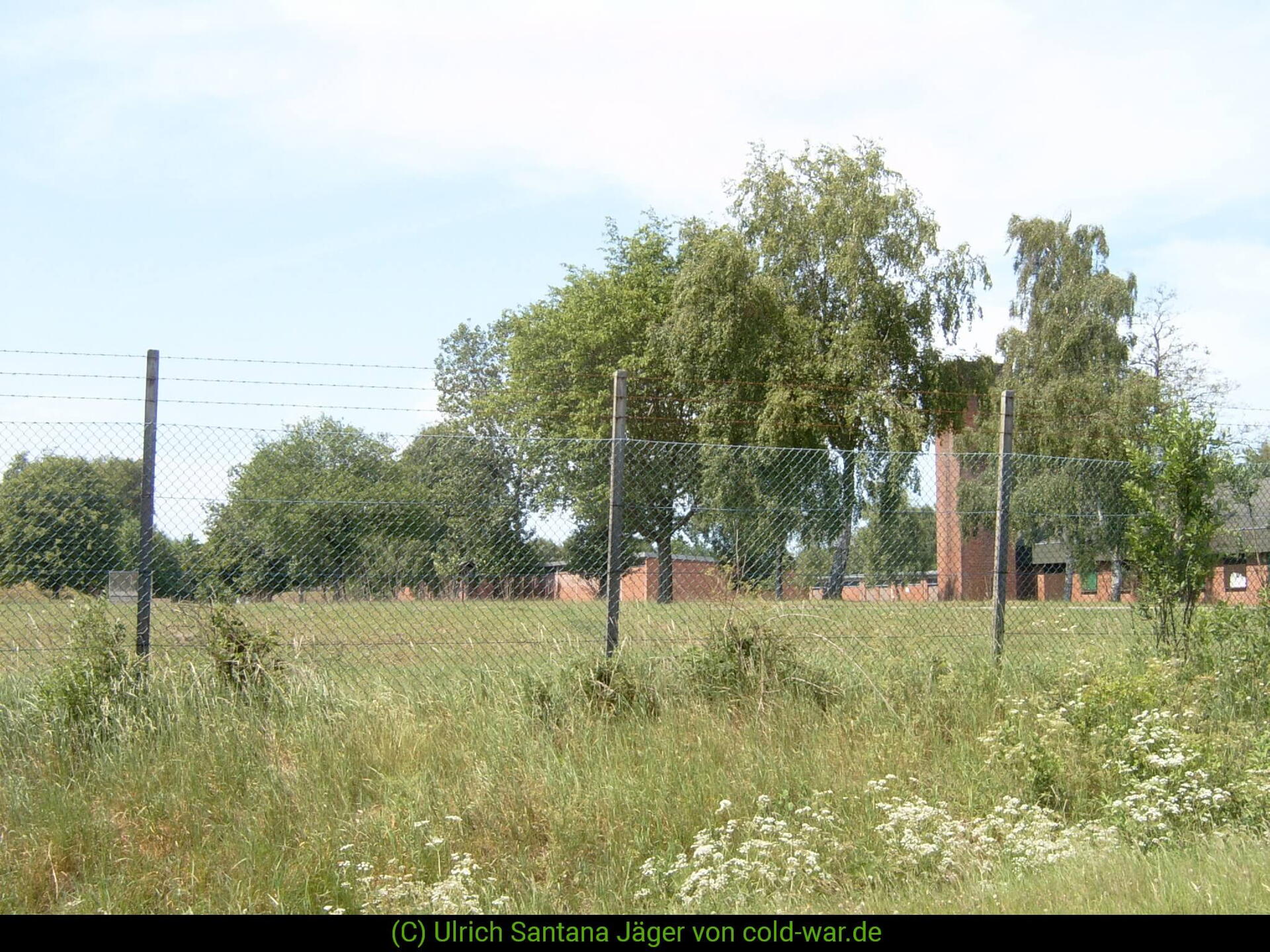 Ein umzäuntes Gelände mit Backsteingebäuden und Bäumen unter blauem Himmel in Idstedt. | © Ulrich Santana Jäger von cold-war.de