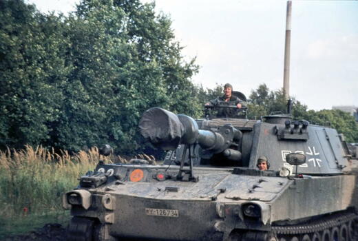 Ein Panzer der Bundeswehr fährt mit Soldaten in der Natur, umgeben von Bäumen und hohem Gras.