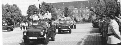 Militärfahrzeuge fahren an salutierenden Soldaten vorbei, während sie vor einem großen Gebäude eine Parade abhalten.
