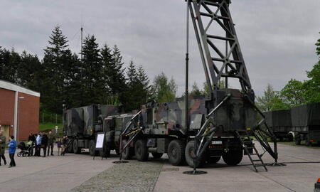 Ein großer Militärtruck mit aufgerichtetem Radarsystem steht auf einem Platz, umgeben von Bäumen und Gebäuden.