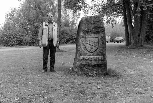 Ein Mann steht neben einem Gedenkstein mit Inschrift und Wappen in einer parkähnlichen Umgebung.