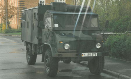 Ein militärischer MB Unimog mit Tarnmuster parkt auf einer regennassen Straße vor einem Backsteingebäude.