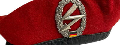 Ein rotes Barett der Fernmeldetruppe mit einem silbernen Emblem und deutscher Flagge ist abgebildet.