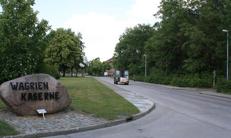 Ein großer Stein mit der Aufschrift "Wagrien Kaserne" steht neben einer ruhigen, von Bäumen gesäumten Straße.
