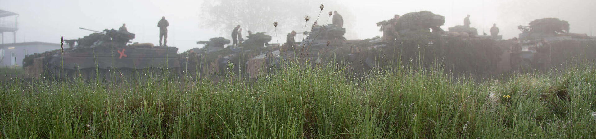 Panzer und Soldaten üben im morgendlichen Nebel hinter einer grünen Wiese während des Gefechtsbild-Trainings.