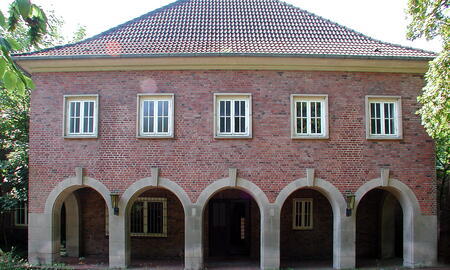 Backsteingebäude mit fünf Bögen und Fenstern im Hauptportal, umgeben von Bäumen an einem sonnigen Tag.