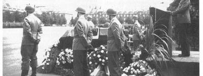 Ein Redner hält eine Ansprache bei einer militärischen Zeremonie neben einem mit Blumen geschmückten Pult, umgeben von Soldaten.