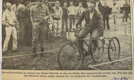 Ein Mann fährt ein modifiziertes Dreirad auf einer Wiese, während eine Gruppe von Zuschauern in Ladelund lacht.