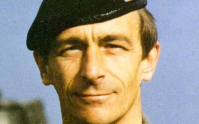Ein Mann in militärischer Uniform und schwarzem Barett steht draußen vor einem blauen Himmel.