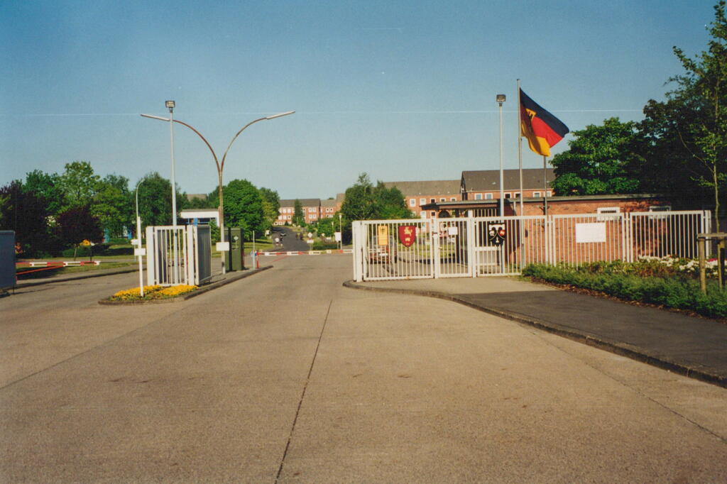 Ein militärischer Eingang mit deutscher Flagge und Wappen, umgeben von Bäumen und einem sonnigen Himmel.