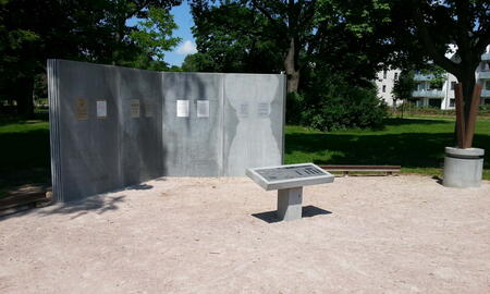 Ein Denkmal mit Informationstafeln steht auf einem sandigen Platz, umgeben von grünen Bäumen und modernen Gebäuden.