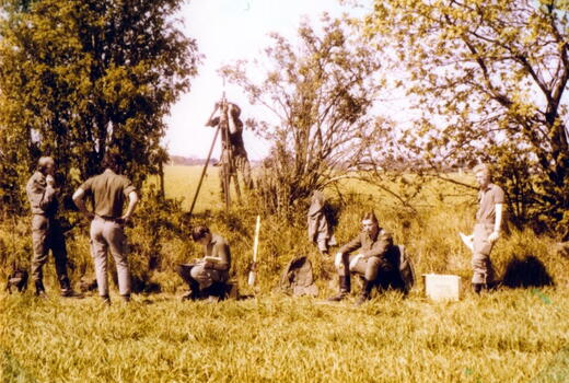 Eine Gruppe von Männern führt Vermessungsarbeiten auf einem Feld durch, umgeben von Bäumen und Gräsern.