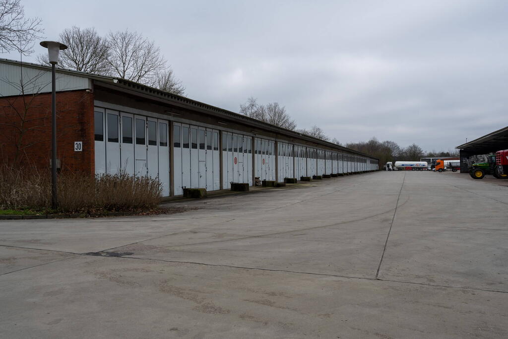 Ein langer, weißer Lagergebäude-Komplex steht neben einem leeren Betonhof unter einem bewölkten Himmel.