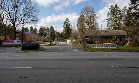 Der Eingang der Waldersee-Kaserne zeigt eine ruhige Straße, umgeben von Bäumen und Backsteingebäuden unter einem bewölkten Himmel.