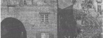 Eine historische Militärparade zieht durch eine Altstadtstraße, vorbei an einem markanten, alten Backsteingebäude.