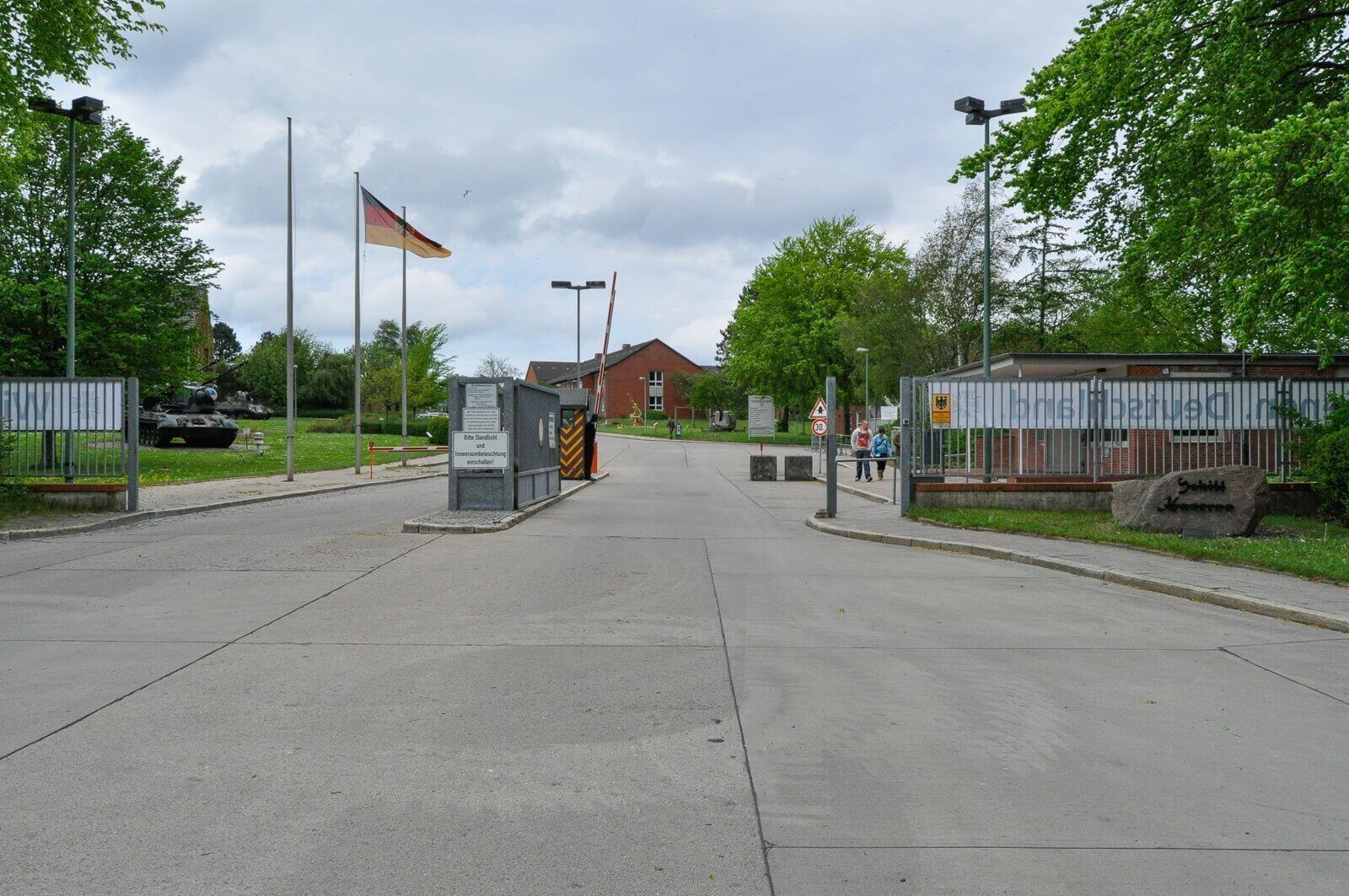 Ein deutscher Militärstützpunkt mit wehender Flagge, einem Panzer links und zwei Personen am Eingangstor.