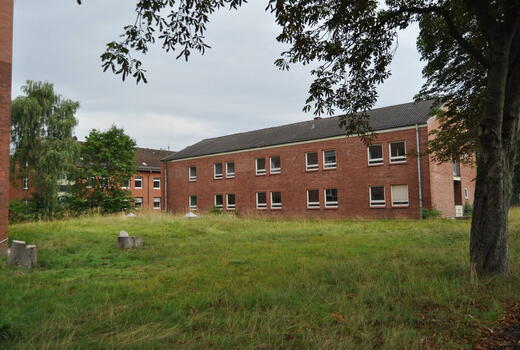 Ein rotes Backsteingebäude der Eider-Kaserne steht inmitten einer bewachsenen Rasenfläche unter bewölktem Himmel.