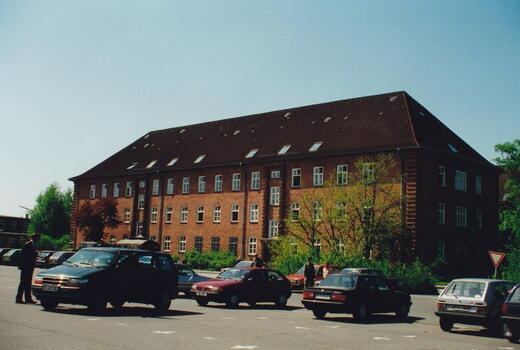 Autos parken vor der Bismarck-Kaserne, einem großen Backsteingebäude mit zahlreichen Fenstern, an einem sonnigen Tag.