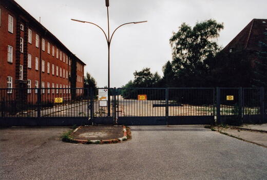 Ein geschlossenes Tor vor der Bismarck-Kaserne von 1996, umgeben von einem Backsteingebäude und Bäumen.