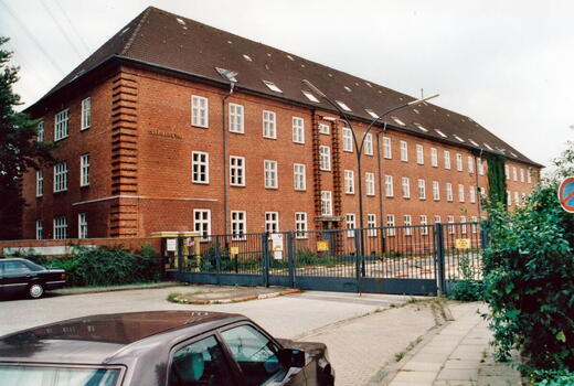 Ein großes Backsteingebäude der Bismarck-Kaserne steht hinter einem geschlossenen Tor, umgeben von parkenden Autos.