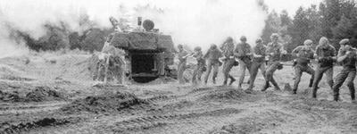 Soldaten ziehen in einer Reihe ein gepanzertes Fahrzeug auf einem staubigen Feld, umgeben von Rauchwolken.