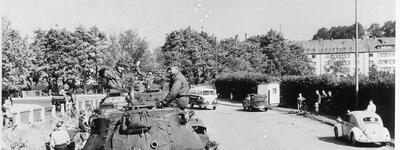 Soldaten auf einem US-Armee-Panzer fahren 1956 durch eine deutsche Straße, flankiert von Autos und neugierigen Fußgängern.