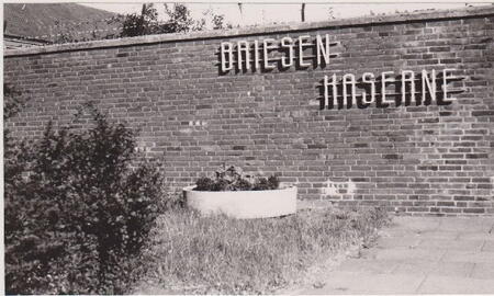 An einer Ziegelmauer steht der Schriftzug "Briesen-Kaserne" über einem Pflanzkübel im Gras.