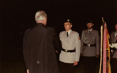 Ein Mann in Uniform wird bei einer nächtlichen Zeremonie von einem älteren Herrn begrüßt, während Fahnen im Hintergrund wehen.