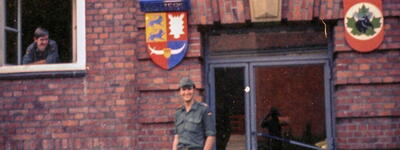 Ein Mann in Uniform steht vor einem Backsteingebäude, während ein anderer aus einem Fenster schaut, mit Wappen an der Wand.