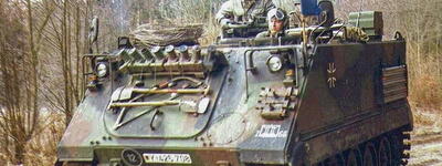 Ein M113-Panzerwagen fährt durch einen bewaldeten Weg, während zwei Soldaten aufmerksam die Umgebung beobachten.