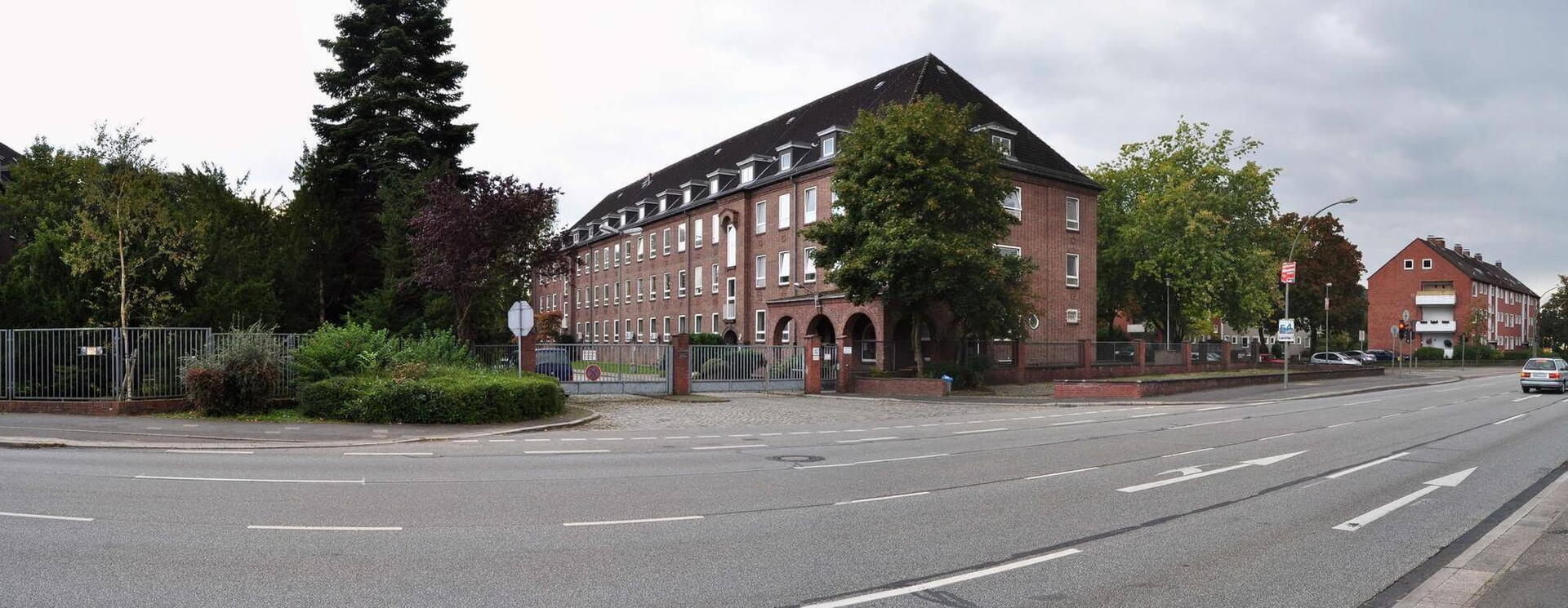 Ein historisches Backsteingebäude der Scholtz-Kaserne von 2008 steht an einer ruhigen Straßeneinfahrt, umgeben von Bäumen.