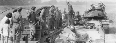 Soldaten und Zivilisten stehen auf Panzern und Lastwagen auf einem Feldweg, während sie miteinander sprechen.