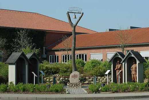 Ein Denkmal mit einer Glocke auf einem Holzgestell steht vor einem Gebäude mit roten Ziegeldächern in Seeth.