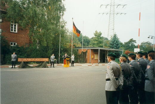 Eine Person hält eine Rede vor Soldaten in Uniformen an der Bismarck-Kaserne, neben einer aufgezogenen deutschen Flagge.
