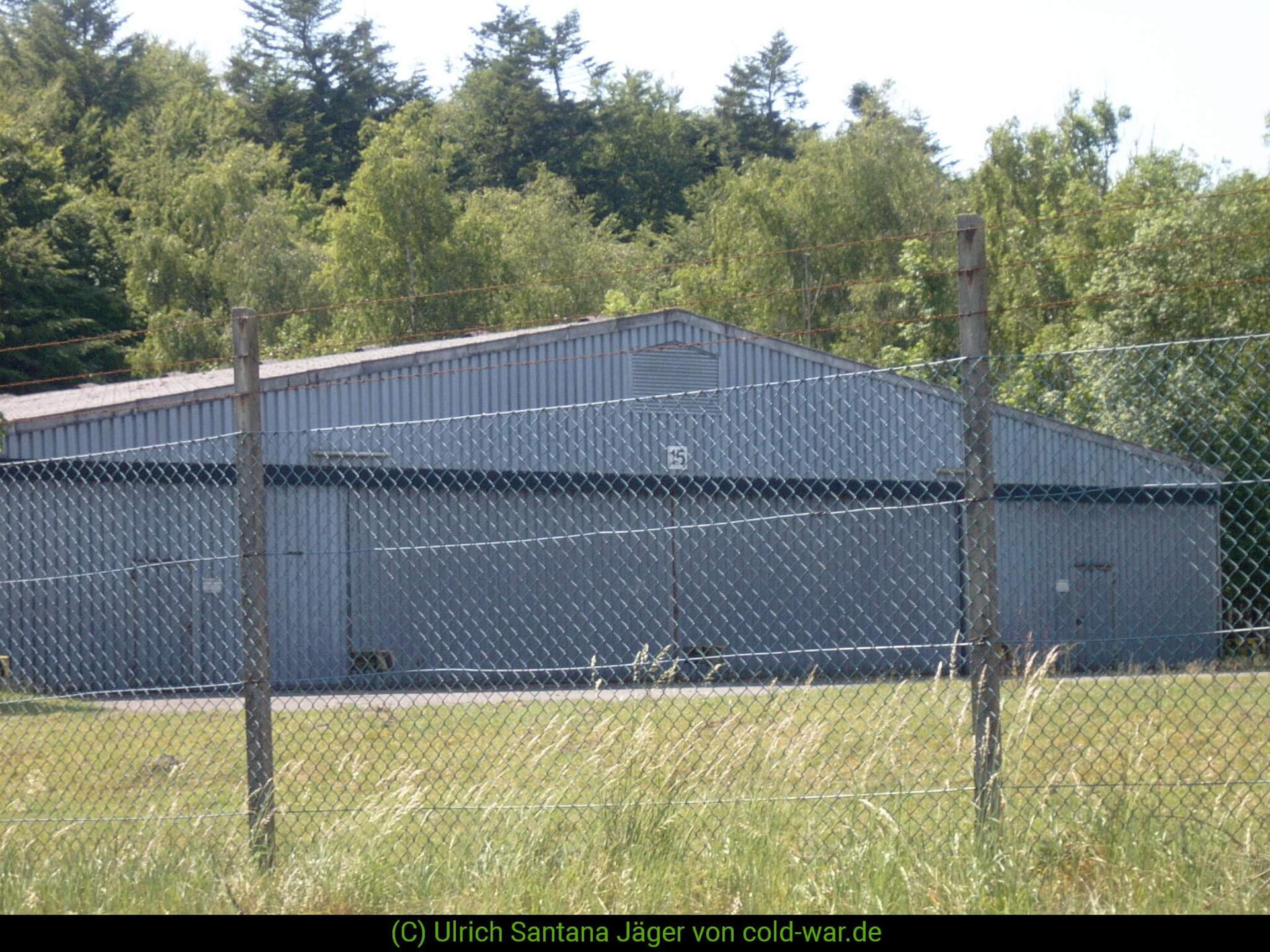 Ein großer, grauer Hangar steht hinter einem Maschendrahtzaun, umgeben von dichter Vegetation. | © Ulrich Santana Jäger von cold-war.de