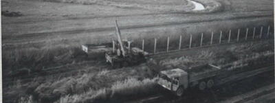 Ein Bagger und ein Lastwagen arbeiten an der Errichtung eines Zauns auf einem weiten Feld neben einem schlängelnden Bach.