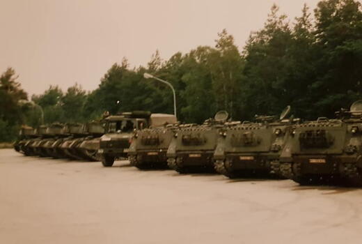 Eine Reihe von Militärfahrzeugen steht nebeneinander auf einem Parkplatz vor einem Wald.