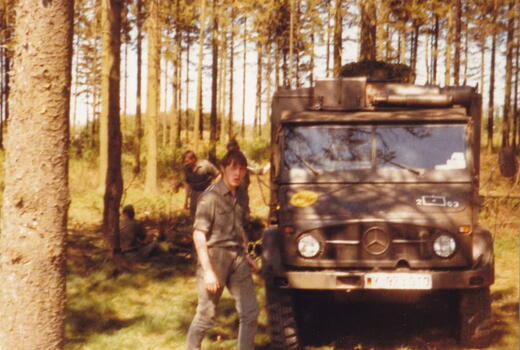 Ein Mann steht neben einem Mercedes-Jeep im Wald, während im Hintergrund weitere Personen zu sehen sind.