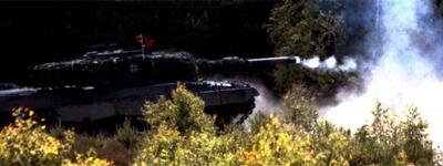 Ein Panzer feuert in einem bewaldeten Gebiet einen Schuss ab, wobei Rauch aufsteigt und Vegetation im Vordergrund zu sehen ist.
