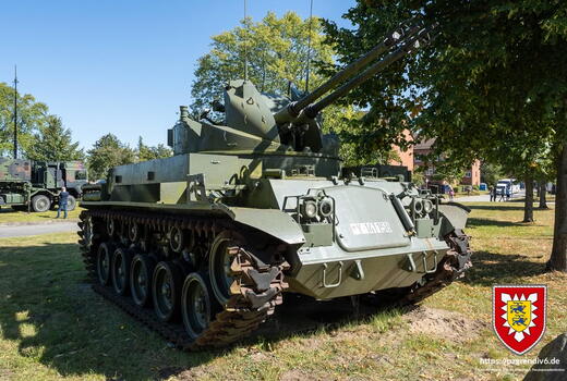 Ein grüner Flakpanzer steht auf einer Wiese in einer Ausstellung, umgeben von Bäumen und Besuchern.
